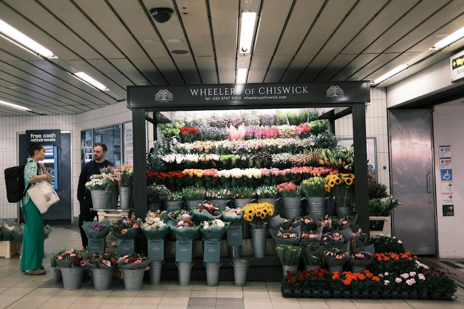 A vibrant floral display from Wheeler of Chiswick within an underground station, featuring a multitude of freshly cut flowers arranged in tiered layers. The arrangement includes yellow sunflowers, whi