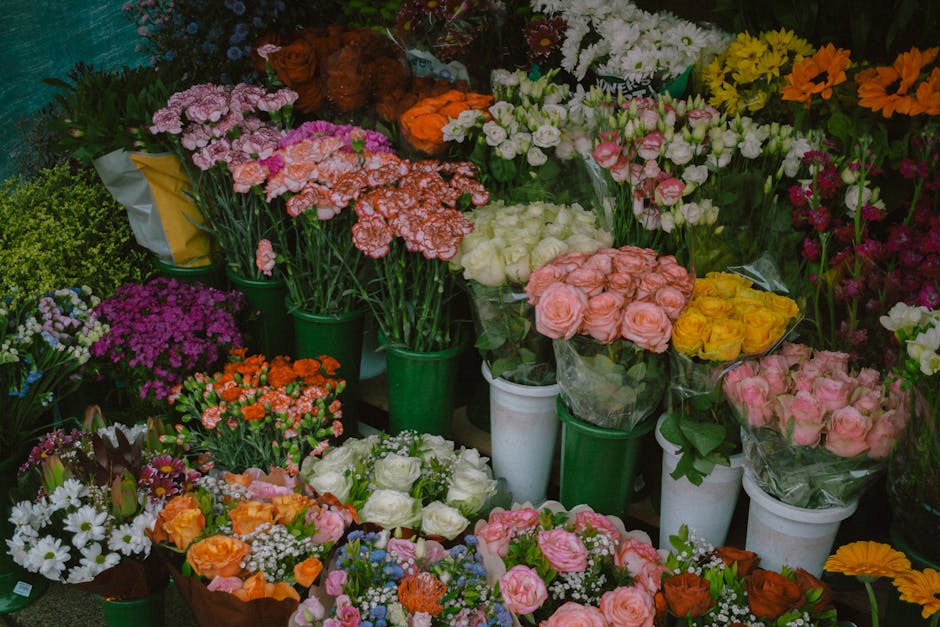 A vibrant array of fresh flowers arranged in various bouquets and vases, including pink roses, white roses, orange carnations, purple chrysanthemums, white daisies, and yellow blooms. The flowers are