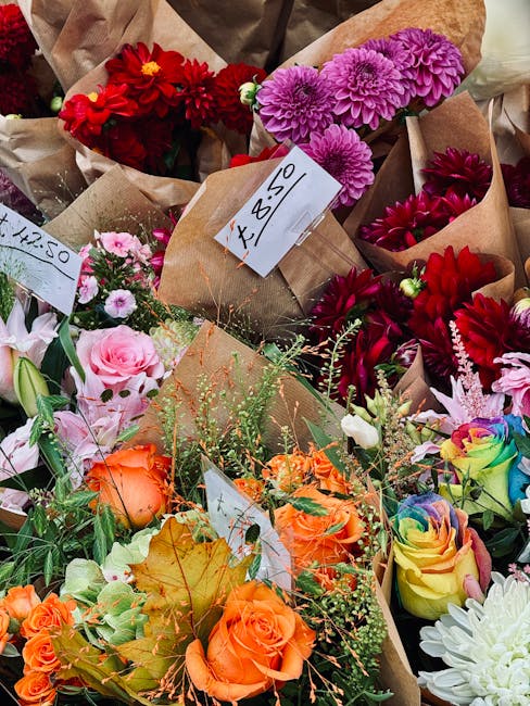 A colorful assortment of fresh flowers arranged in small brown paper wrapping, including vibrant red dahlias, purple chrysanthemums, pink roses, orange roses, and multicolored roses. The bouquets are 