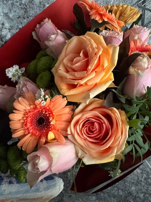A bouquet of fresh flowers arranged with peach and light pink roses, orange gerbera daisies, and pale pink lisianthus, complemented by green foliage and small white filler flowers. The flowers are wra