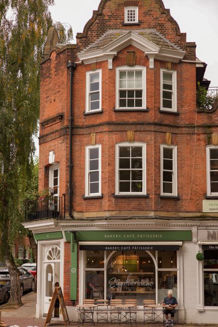The image depicts a charming red-brick building with a bakery, café, and pâtisserie on the ground floor, featuring large display windows and a green awning. Above, the upper floors include white-fra