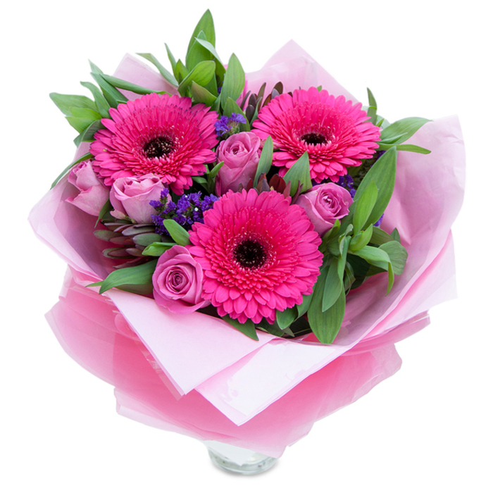 The charm here is immediate: three magnificent fuchsia Gerberas stand proud at the heart of the design, their intense pink petals radiating around dark, textured centres that anchor the arrangement with confident contrast. Surrounding them are roses in a tender lavender-pink or dusty rose, their petals still compact and fresh, suggesting both delicacy and the promise of fuller bloom. Scattered between these principal flowers are small sprays of purple statice, adding a dry, papery texture and a cooler violet note that refracts the pinks into a softer chorus. Generous green foliage-salal leaves with broad, glossy faces and a suggestion of eucalyptus-creates volume and frames each bloom, offering a crisp, slightly dewy green scent that complements the floral perfume. The bouquet nestles in an inner sheath of matte pale-pink tissue paper, while an outer wrap of translucent fuchsia-pink cellophane, gently crinkled, adds a filmy glint and continuity of tone; the wrapping is folded with artisanal precision, and the top lip of a clear glass vase peeks out at the bottom, indicating the bouquet is ready to display. Set against a pristine white backdrop and lit with soft, even light, the textures-velvet petals, papery statice, glossy leaves-are easy to read and palpably fresh. This arrangement carries a bright, affectionate energy, perfect for marking a birthday, an anniversary, or a thoughtful 'just because' gesture after an afternoon stroll through Sheen Common or a coffee on Upper Richmond Road, bringing immediate cheer and a tactile sense of care to whoever receives it.
