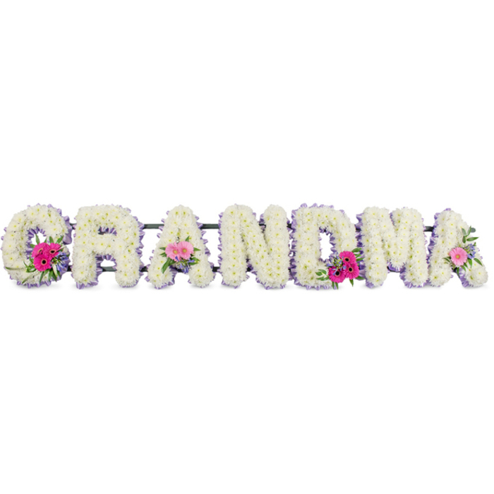 In this touching floral display, the word GRANDMA stretches in a row of substantial letters, each one deeply cushioned and densely packed with white chrysanthemums. The flowers form a clean, uninterrupted surface, so the letters look smooth and pillowy, their rounded petals hinting at softness and comfort. Encircling each letter is a delicate lilac ribbon, carefully attached so that its edge follows the shape of the letter without breaking the neat line of blooms. The ribbon adds a faint glow of colour-a gentle pastel purple that symbolises grace and tenderness-and subtly separates the white flowers from the background. Three letters carry extra detail: the G at the beginning, the A at the centre, and the final A. On these, small hand-crafted floral sprays are pinned into place, made of bright pink gerbera daisies with bold, radiating petals, romantic pink roses just opening, and mixed green foliage. The greenery ranges from soft, feathery textures to glossy leaves, creating contrast against the chrysanthemums. These posies sit like little islands of colour on the white sea of petals, suggesting moments of joy and individual stories within a shared family history. The tribute is photographed against a plain backdrop in soft, diffused light, which ensures the whites remain clear and the pinks appear natural and true. The piece feels like a thoughtful farewell for a grandmother in East Sheen, perhaps chosen for a service at a local church off Upper Richmond Road or for delivery to a funeral director along Sheen Lane, expressing respect, gratitude, and enduring affection through carefully chosen flowers.